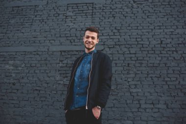 Kot gömlekli, siyah ceketli, elini cebinde tutan ve siyah tuğlalı duvarın yanındaki kameraya bakan neşeli bir genç.