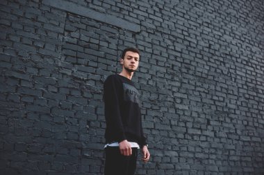 Sokakta yüksek koyu tuğlalı duvarın yanında durmuş kameraya bakan, modaya uygun giyinmiş, ciddi, genç bir etnik erkek.