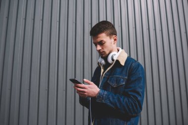 Kot ceketli, kulaklıklı, cep telefonlu, sokakta metal çitlerin yanında duran ciddi bir gencin yan görüntüsü.