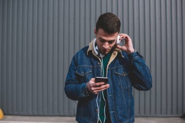 Kulaklıklı ve kot ceketli adam metal çitin yanındaki sokakta müzik dinlerken akıllı telefon ekranına bakıyor.