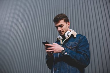 Düşük açılı, sıradan ceketli, kulaklıklı, kulaklıklı, metal çitlerin yanındaki akıllı telefondan internette gezinen bir adam.