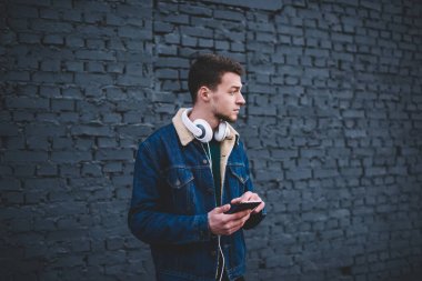 Sıradan giyinen, kulaklık takan, akıllı telefonlara bakan ve şehir sokağındaki tuğla duvara bakan düşünceli bir adam.