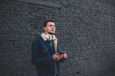Kot ceketli, kulaklıklı, kulağında kulaklıkla düşünceli bir adam. Şehir caddesinde, tuğla duvarın yanındaki akıllı telefona bakarken dikkatle başka yere bakıyor.