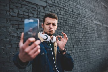 Kot ceketli ve kulaklıklı genç, sakallı, etnik bir erkek. Göz kırparak selfie çekiyor.