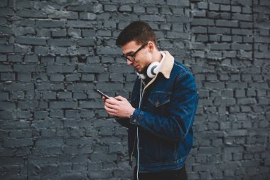 Kot ceketli ve kulaklıklı gülümseyen genç adamın yan görüntüsü sokakta tuğla duvarın yanındaki cep telefonuyla mesaj gönderiyor.