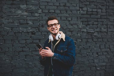Moda kıyafetleri içinde, kulaklıklı, cep telefonlu, neşeli genç bir adam sokakta tuğla duvarın yanında durup kameraya bakıyor.