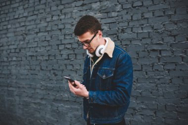 Kot ceketli, kulaklıklı, cep telefonlu, siyah tuğlalı duvarın yanında duran genç adamın yan görüntüsü.