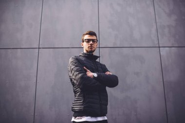 Modern binanın yanında dikilip kameraya bakan sıradan giysili ve gözlüklü havalı genç bir erkek.