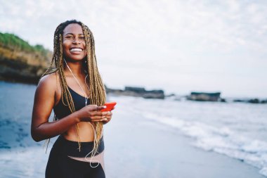 Kulaklıkla kulaklıkla müzik dinlemekten zevk alan mutlu Afrikalı Amerikalı koşucu, cep telefonundan müzik dinlemek için aktif kıyafet giymiş komik koyu tenli koşucu.