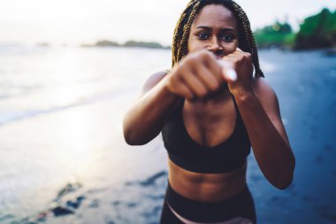 Aktif giyinen Afrikalı Amerikalı sporcu kızın yarım boy portresi deniz kıyısındaki spor eğitiminde ellerin şeklini geliştirmek için boks egzersizleri yapıyor. Dayanıklılık kavramı.