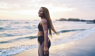 Neşe saçan Afrikalı Amerikalı kadın sabah antrenmanında mutlu oluyor. Başka tarafa bakıyor ve gülümsüyor. Komik koyu tenli, sportif kıyafetler giymiş, deniz kıyısına gülüyor.