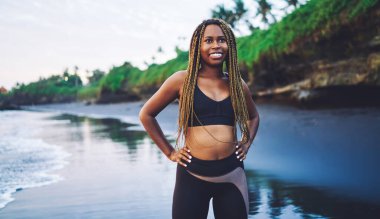 Deniz kıyısında duran ve gülümseyen, eşofman giymiş, spor yapmak için giyinmiş, sağlıklı yaşam tarzının ve canlılığının tadını çıkaran neşeli Afro-Amerikan spor kadını.