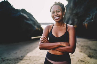 Eşofmanlı, neşeli Afro-Amerikan genç bir kadının portresi kameraya gülümserken elleri çapraz, sportif bluz giymiş, sabah sahile poz vermek için koşan neşeli kadın koşucu.