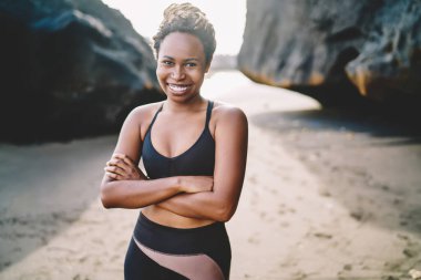Eşofmanlı, neşeli Afro-Amerikan genç bir kadının portresi kameraya gülümserken elleri çapraz, sportif bluz giymiş, sabah sahile poz vermek için koşan neşeli kadın koşucu.