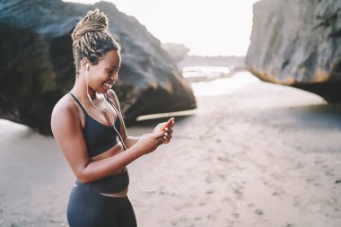 Neşeli koyu tenli kadın koşucu, sahilde sabah antrenmanı için radyoda müzik seçerken mutlu hissediyor. Elektronik kulaklıklı mutlu bir kadın cep telefonuyla cep telefonuna bağlı bir oyuncunun sesini dinliyor.