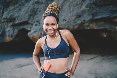 Cep telefonunda müzik uygulamasından gelen ses şarkılarını dinlerken gülümseyen neşeli Afro-Amerikan kadın portresi. Sportif bluz giymiş komik esmer koşucu sabah keyfine bakıyor.