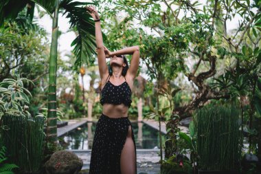 Yeşil Park 'ta, güneşli yaz gününde tropikal tatil köyünde dans eden ve el kaldıran modaya uygun giyinmiş sıska genç kadın.