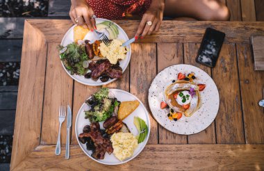 Yüksek açılı, yüzü olmayan, iştah açıcı bir kahvaltı yapan bir kadın omlet, haşlanmış patates ve ahşap masaya yerleştirilmiş domuz pastırması ve açık kafedeki tatlı gözlemeler.