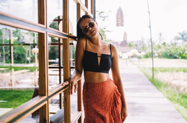度假期间 身穿时髦夏装 戴着太阳镜 靠着现代建筑的玻璃墙 身穿浅褐色日光浴的年轻女性旅行者 — 图库照片
