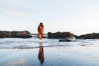 Genç, uzun saçlı, turuncu elbiseli, Bali 'de parlak bir günde dalgalı deniz kıyılarında gezinen, uzun boylu, etnik bir kadın.