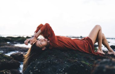 Kırmızı cüppeli, elleri kafasında, bacakları bükülmüş ve dinlenirken kameraya bakan zarif esmer kadın.