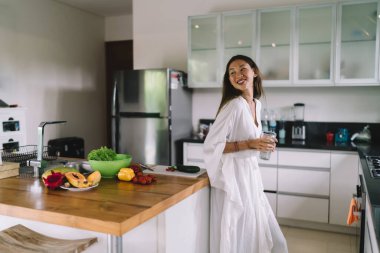 Mutfakta bir bardak su içerken masaya yaslanmış egzotik meyve ve sebzelerle birlikte beyaz cüppeli muhteşem etnik kadın manzarası.