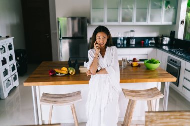 Beyaz bornozlu zarif bir Asyalı kadın. Tezgaha eğilip modern mutfakta kahvaltı yaparken kameraya bakıyor.