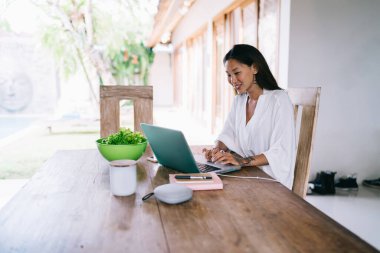 Güler yüzlü Asyalı serbest yazar, şık beyaz elbisesiyle, tahta masada dizüstü bilgisayarında, gündüz vakti hafif ferah bir odada sağlıklı salatayla geziyor.