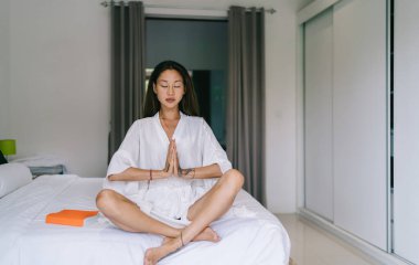 Yoga pozunda gündüz vakti yatak odasında bacak bacak bacak üstüne atmış şekilde otururken gözleri kapalı, sakin Asyalı kadın el ele tutuşuyor.