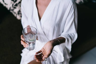 Yüksek açılı, tanınmayan, bornozlu bir kadın rahat koltukta oturuyor ve Bali 'de hap içerken bir bardak su içiyor.