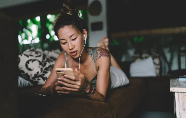 Татуированная Азиатка Лежит Диване Лежа Животе Используя Смартфон Наушники Уютной — стоковое фото