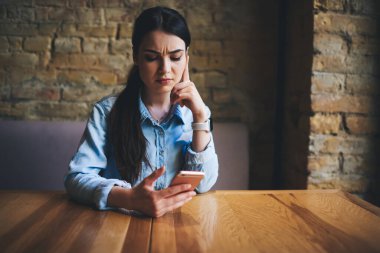 Tatminsiz uzun saçlı kadın ahşap masaya yaslanmış ve dikkatle ekrana bakarken çağdaş kafede duvarları tuğladan duvarlarla çevrili akıllı telefona bakıyor.
