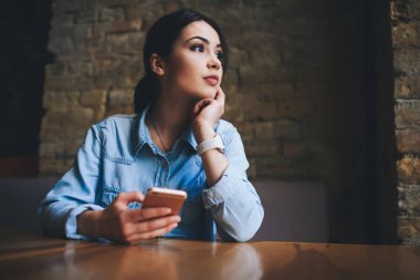 Modern kafede arka planında tuğla duvar olan akıllı telefonlara bakarken gündelik giyinmiş, düşük açılı bir kadın.