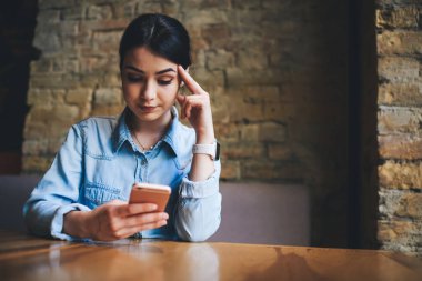 Düşük açılı, ahşap masada oturan ve arka planda tuğla duvar olan modern kafede akıllı telefon kullanan kadın.
