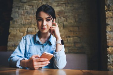 Sıradan giyinmiş hoş bir kadının modern bir kafede elinde akıllı telefonuyla kameraya bakması ve arka planda tuğla duvarla durması.