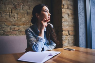 Modern kafede defter yazarken, elinde eğilip yüzünü çeviren, gündelik giysili, dalgın, koyu renk saçlı kadının yan görünüşü.