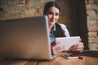 Genç pozitif kadın serbest çalışan kameraya bakarken açık dizüstü bilgisayarla masaya oturuyor ve tablet kullanarak proje üzerinde çalışıyor.
