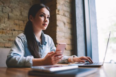 Düşük açılı kadın uzaktan kumandalı sıradan bir işçi akıllı telefon kullanırken ve dizüstü bilgisayarda yazarken masaya bakıyor.