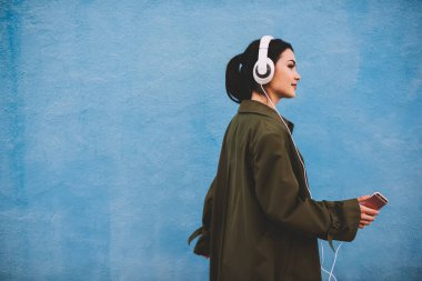 Sıradan giysiler içinde genç bir kadının akıllı telefonuyla yürürken ve kulaklıkla müzik dinlerken yan görüntüsü.