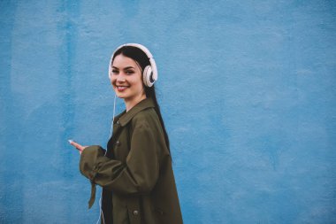 Siyah saçlı, kameraya bakan ve kulaklıklı, mavi arka planda müzik dinleyen neşeli genç bayanın yan görüntüsü.