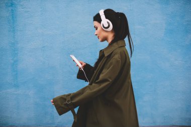 Moda kıyafetleri içinde sakin bir kadın cep telefonuyla gezerken ve kulaklıkla müzik dinlerken yan görünümü