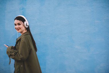 Modaya uygun giyinen, siyah saçlı, gülümseyen ve kameraya bakan, kulaklıklı, mavi arka planda müzik dinleyen neşeli bir kadın.