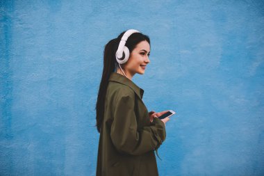 Şık ceketli, akıllı telefon kullanan, kulaklıkla müzik dinleyen ve mavi arka plana bakan bir kadının yan görüntüsü.