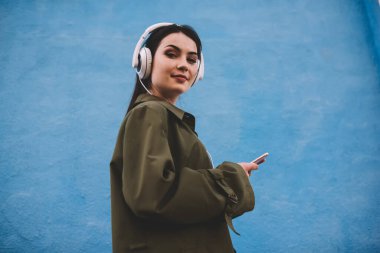 Düşük açılı, siyah saçlı, cep telefonu kullanan ve kulaklıkla mavi duvara karşı müzik dinleyen neşeli bir kadın.
