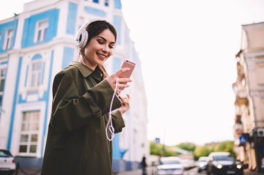 Dişli, güler yüzlü, sıradan elbiseli bir kadın akıllı telefonuyla caddede duruyor ve ekrana bakarken kulaklıkla müzik dinliyor.