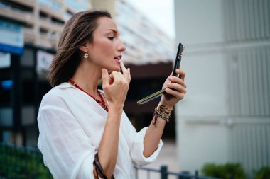 Beyaz tenli kadın turist dudak bakımı için makyaj kremi kullanıyor. Şehir ortamında dikiliyor ve ayna ile palet kullanıyor. Aynaya bakan çekici kadın.