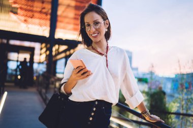 Elinde cep telefonuyla kameraya gülümseyen neşeli kadın blogcunun yarım boy portresi, dışarıda akıllı telefon teknolojisi kullanarak görüş koruması için optik gözlüklü mutlu beyaz kadın.