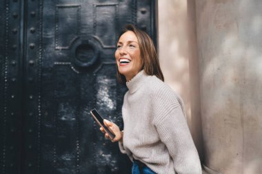 Normal giysiler içinde neşeli yetişkin bir kadın eski binanın girişinde dikilirken gülümsüyor ve akıllı telefonlar çalıyor.