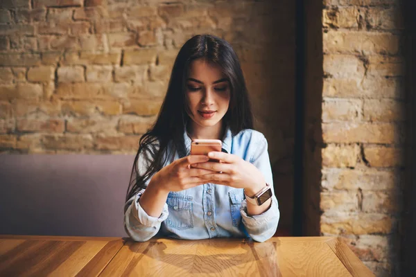 在现代咖啡馆度过周末的时候 穿着休闲装的黑发女士靠着手肘坐在木制桌子上 用智能手机发短信 — 图库照片
