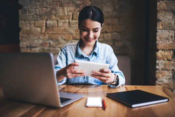 Mutlu kadın serbest çalışan, dişlek gülüşlü modern dizüstü bilgisayarlı ve akıllı telefonlu masada oturmuş tableti karıştırıyor ve çalışıyor.